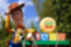 Giant cowboy figure tips hat beside "Toy Story Land" sign made of colorful blocks, set against green foliage and blue sky.