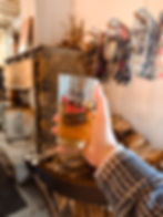 Hand holding a glass of cider labeled "JMASH CIDERY" in a cozy room with a stone fireplace, woodpile, and hanging snowshoes, creating a warm mood.