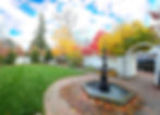 Garden with a black fountain, colorful autumn trees, and a brick pathway. White fence and archway in the background, blue sky above.