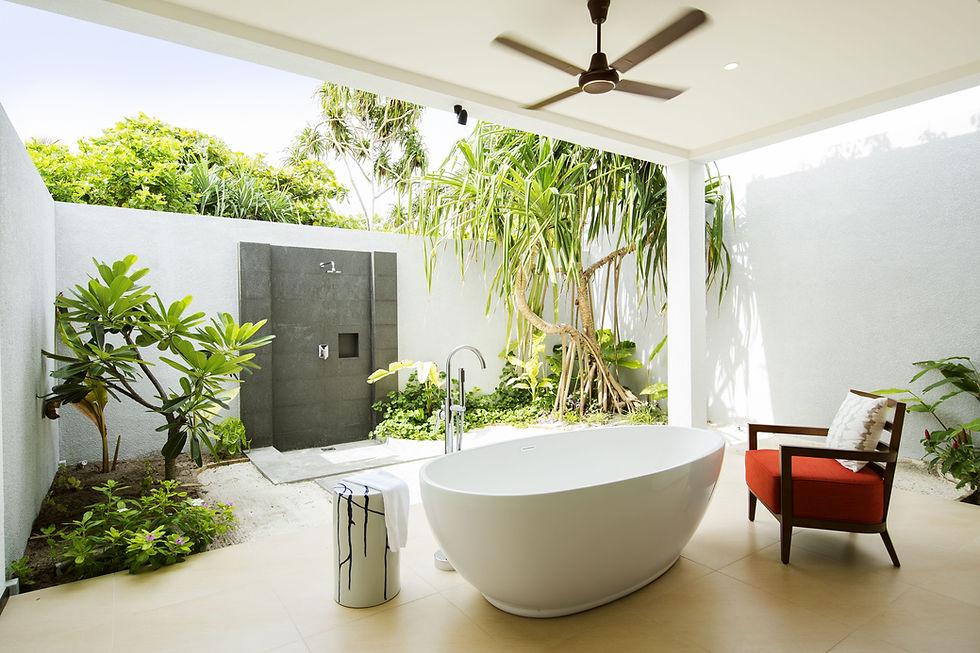 Outdoor bathroom with a white tub, red chair, and black shower against a lush green garden. Ceiling fan and serene ambiance.