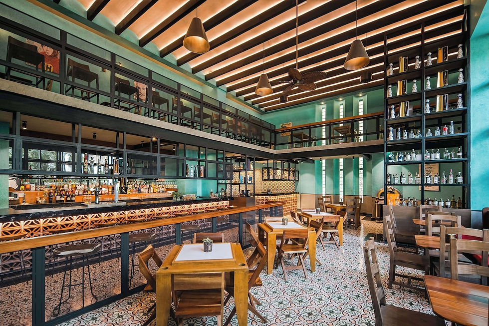 Cozy restaurant interior with wooden tables, chairs, and a bar. Teal walls, patterned floor tiles, hanging lights, and bottles on shelves.