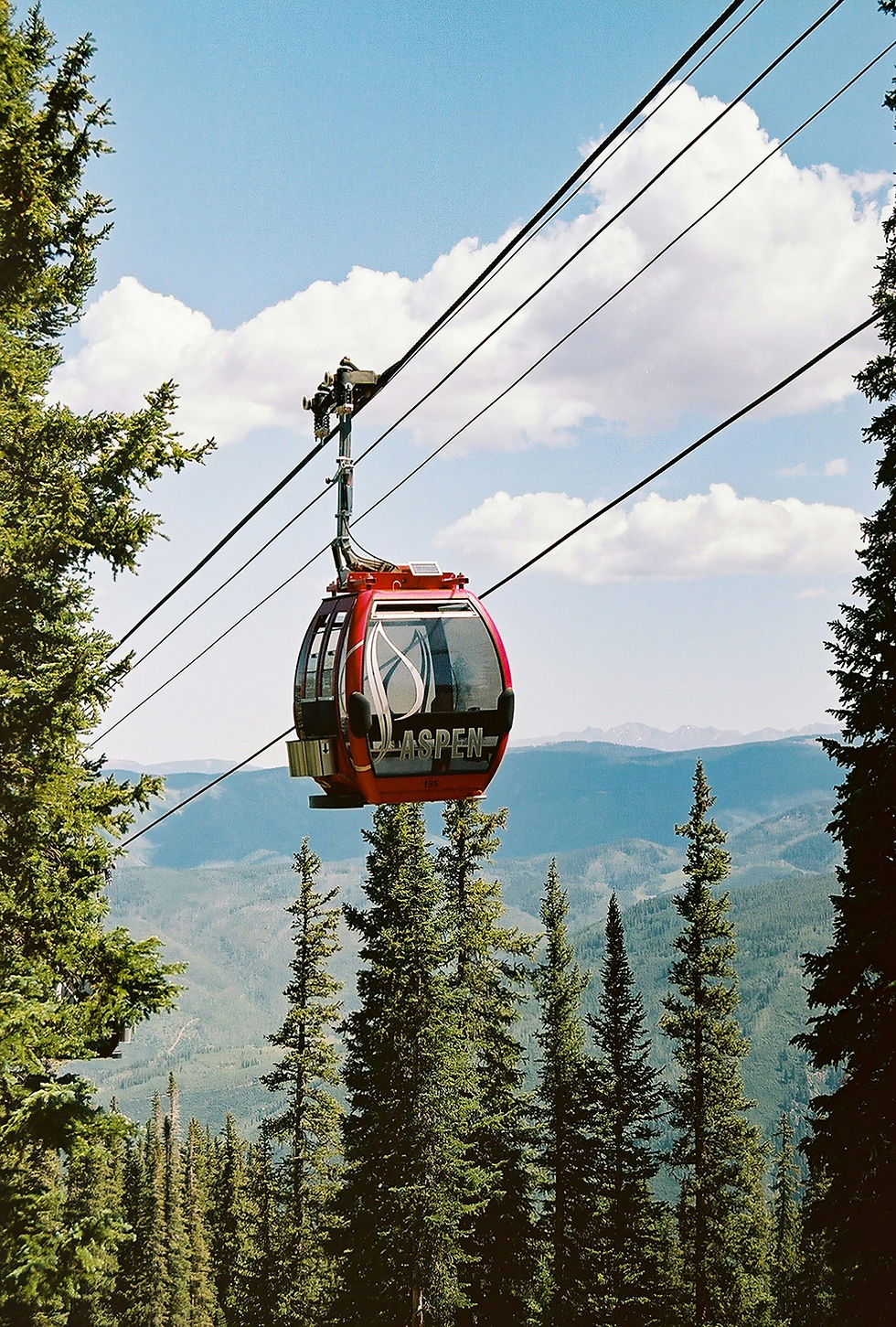 Red gondola marked "Aspen" gliding over lush evergreen trees and mountains under a clear blue sky with fluffy clouds.