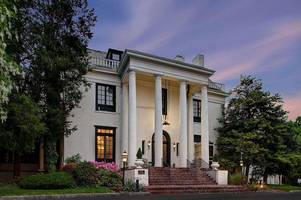 Elegant white mansion with grand columns, surrounded by trees and flowers under a twilight sky. Lit windows add a warm glow.