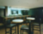 Modern bar with round tables, black chairs, and a countertop lined with bottles. A large black-and-white photo decorates the wall.