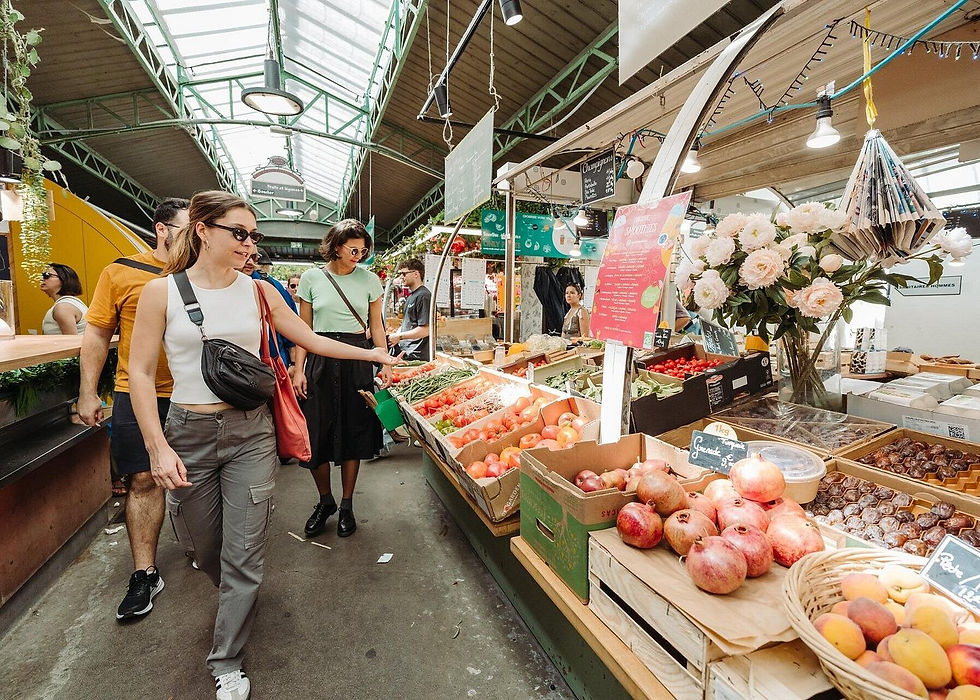 Shoppers browse colorful produce and flowers at a lively indoor market under skylights, with handwritten price signs.