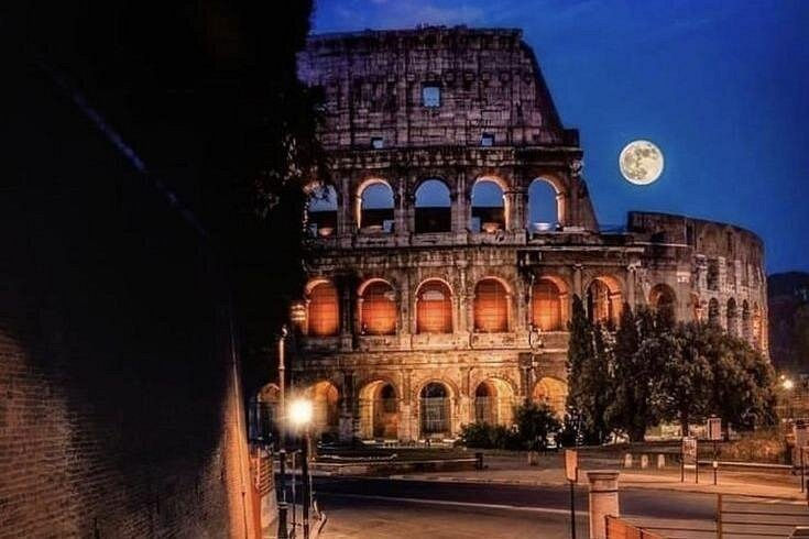 Illuminated Colosseum at night under a full moon, with glowing arches and quiet streets in the foreground.