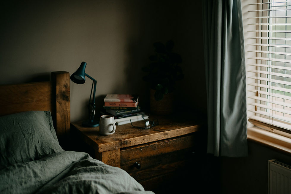 A cozy, rustic bedroom featuring a wooden bedside table with a lamp, books, a ceramic mug, and a pair of glasses. The bed is covered with soft, dark-colored sheets, and natural light streams through blinds partially closed on the window