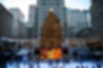 Skaters glide on an ice rink beneath a large, lit Christmas tree. Surrounded by tall buildings, the festive scene features a gold statue.