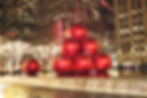 Giant red ornaments stacked near a lit fountain with glowing trees in a cityscape. "Radio City" sign visible on tall buildings.