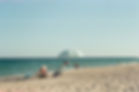 Beach scene with striped umbrella, chairs, and towels. People play by the sea. Bright, sunny day with blue sky and ocean backdrop.