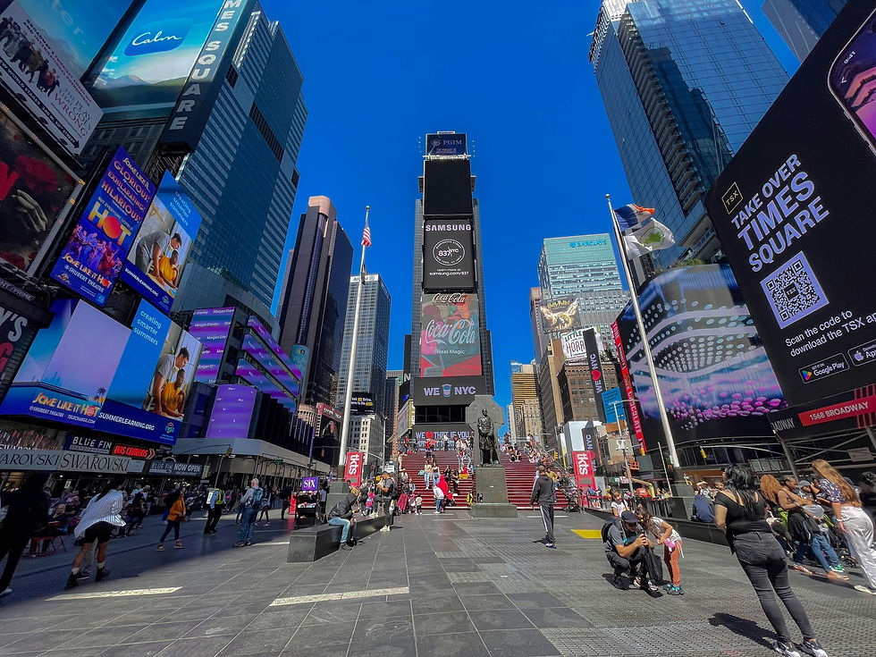 People walk and take photos in Times Square, NYC, surrounded by bright digital billboards. Clear blue sky and vibrant city atmosphere.