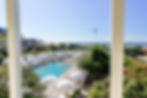 Poolside scene with white umbrellas and loungers, greenery, buildings, and ocean view. U.S. flag visible, creating a tranquil mood.