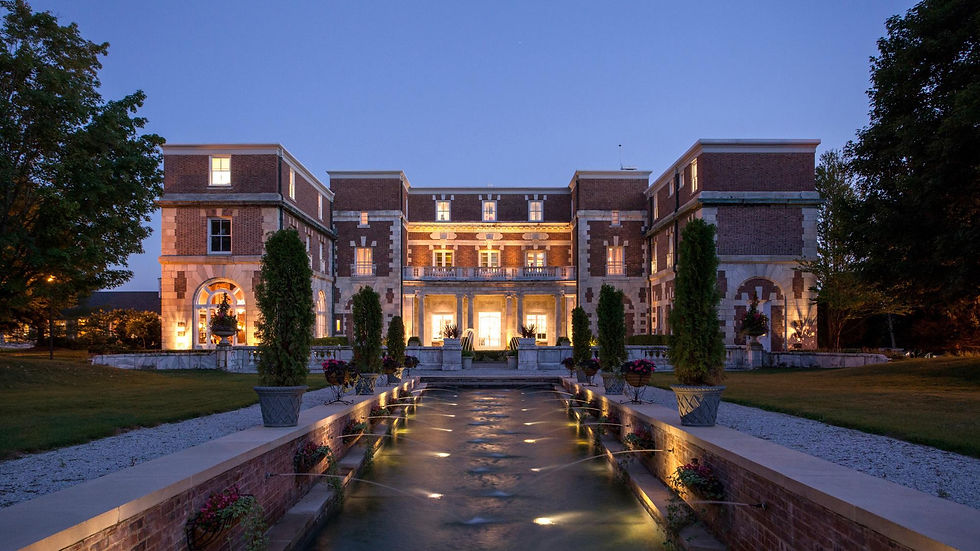 Elegant brick mansion at dusk with glowing lights, fountain in foreground, and manicured trees, set against a clear evening sky.