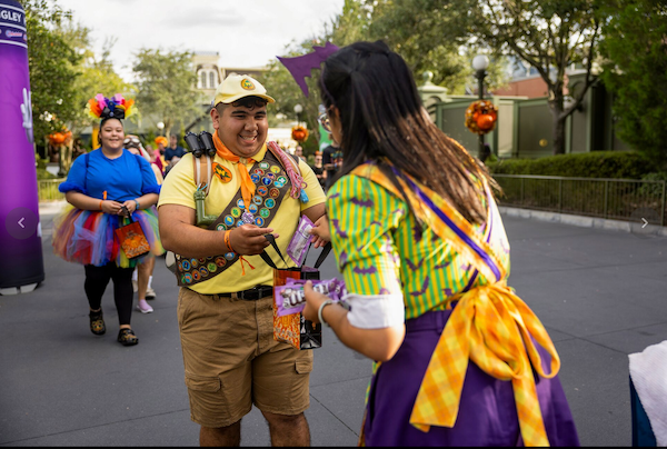 Man in scout costume and woman in colorful attire exchange candy at a park decorated for Halloween. Another person approaches, smiling.