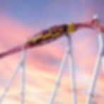Roller coaster with riders on a red track against a vibrant sunset sky. The mood is thrilling and exhilarating as they speed along.