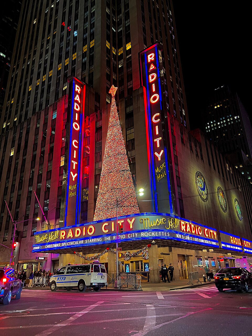 Radio City Music Hall illuminated at night with a Christmas tree decoration above its marquee.
