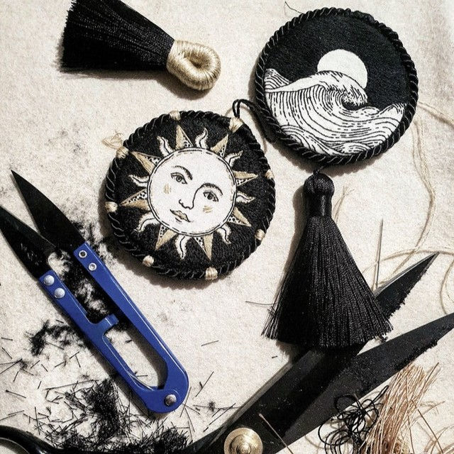 Two embroidered ornaments in progress, with scissors and thread and tassels. Both are circular, and one featured a gold and black sun design, and the other a moon and waves design in black work.