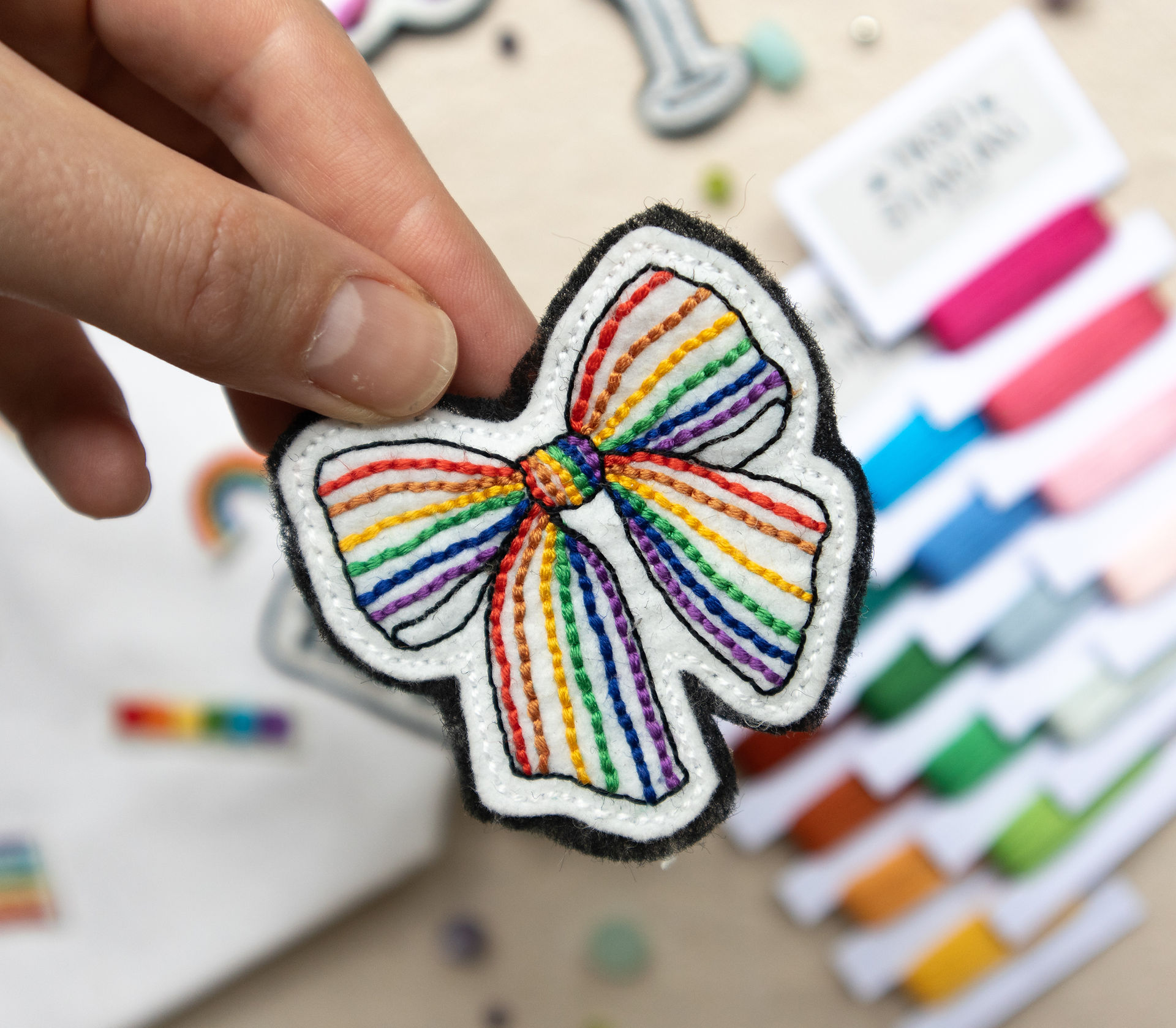 A hand embroidered patches in the shape of a bow, with pride coloured stripes. This is a DIY embroidery patch that is being made at a queer craft workshop in London.
