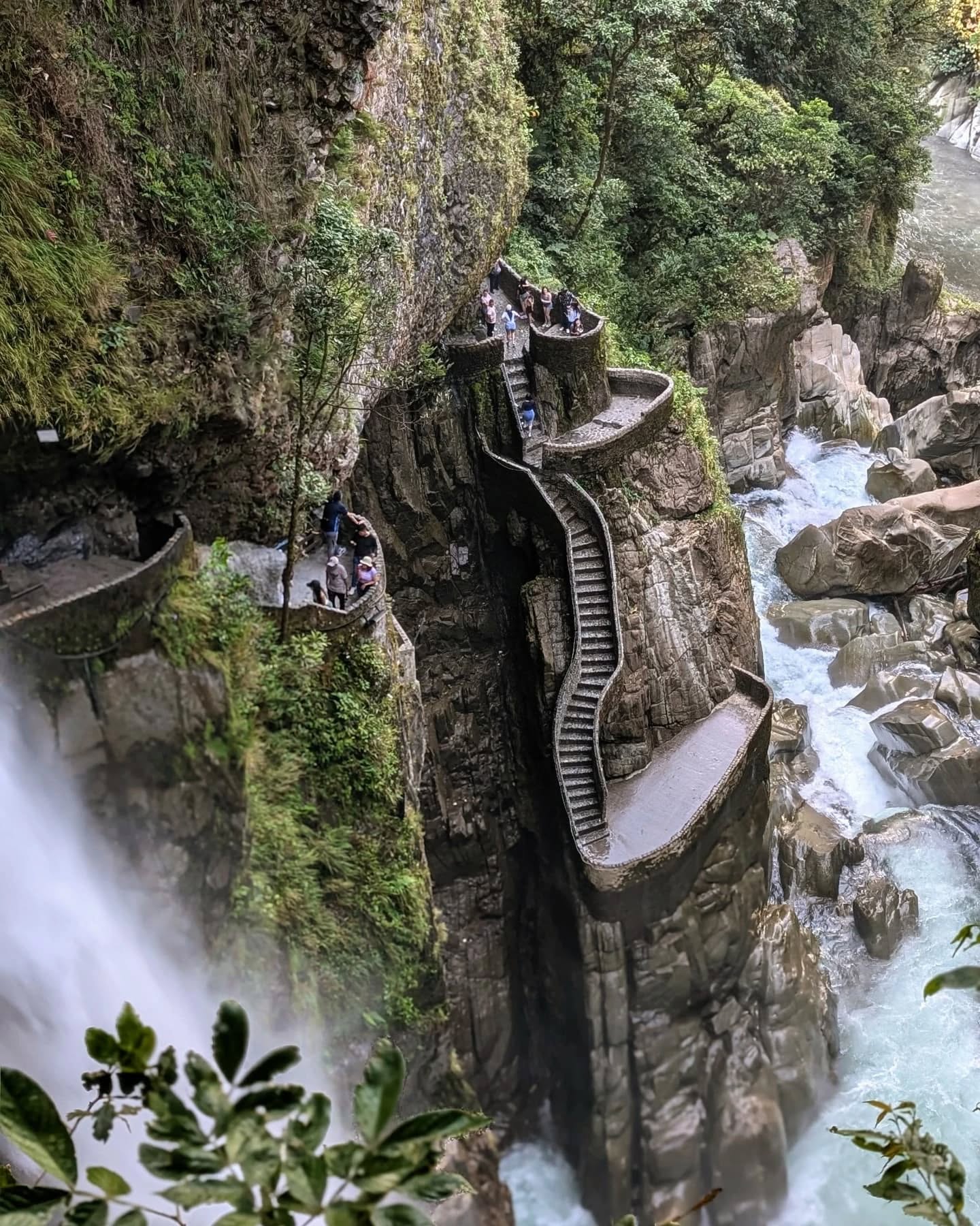 A dramatic view of a steep, winding stone staircase carved into a rocky cliffside next to a powerful waterfall. Visitors walk along the stairs, surrounded by lush greenery and rushing water below. Banos in Ecuador.