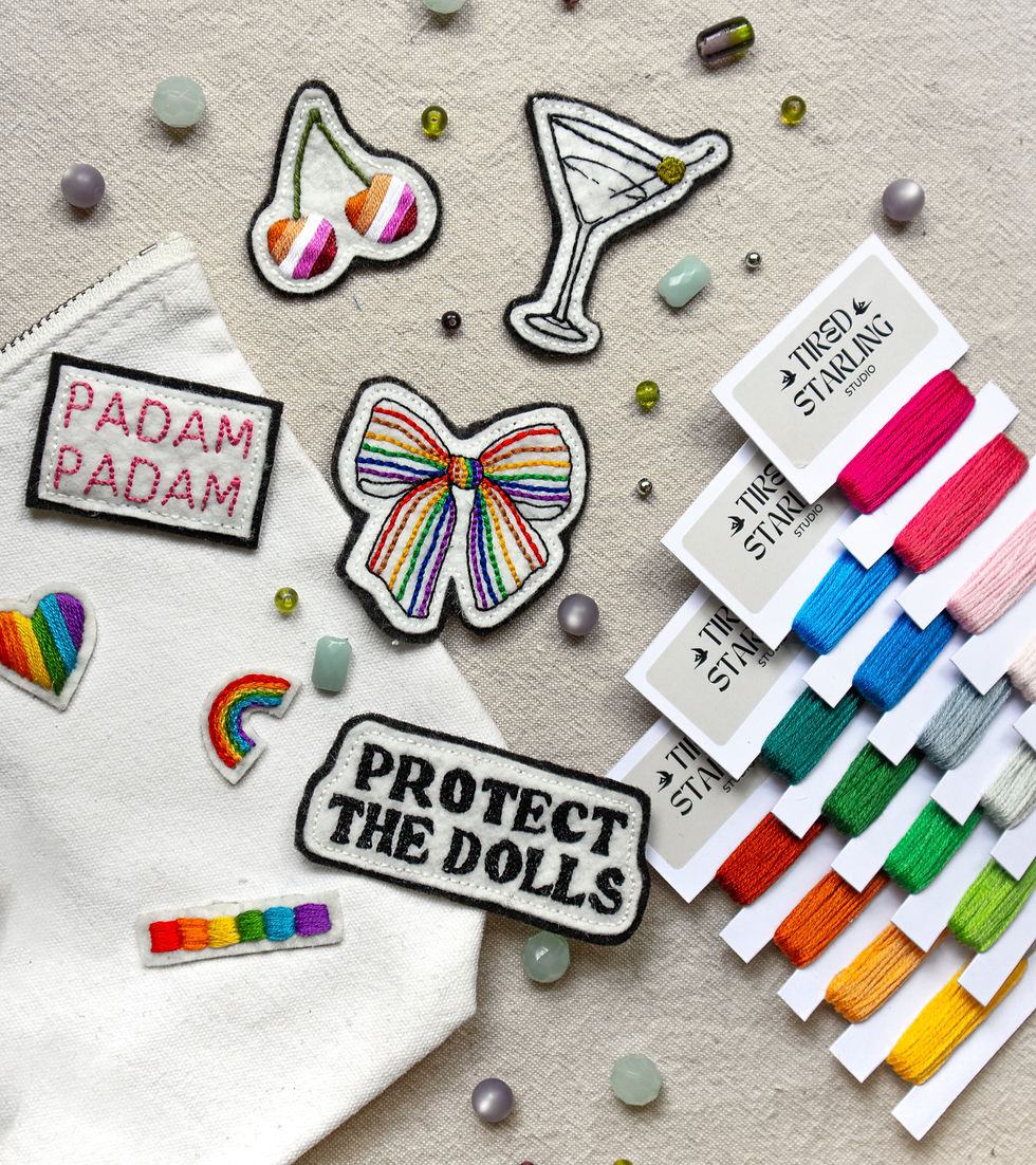 A flat lay photograph of hand embroidered patches for a Queer Embroidery Workshop in London. The patches include cherries, a martini glass, ‘Padam Padam’, ‘Protect the Dolls’, and a bow. All in Pride rainbow colours.