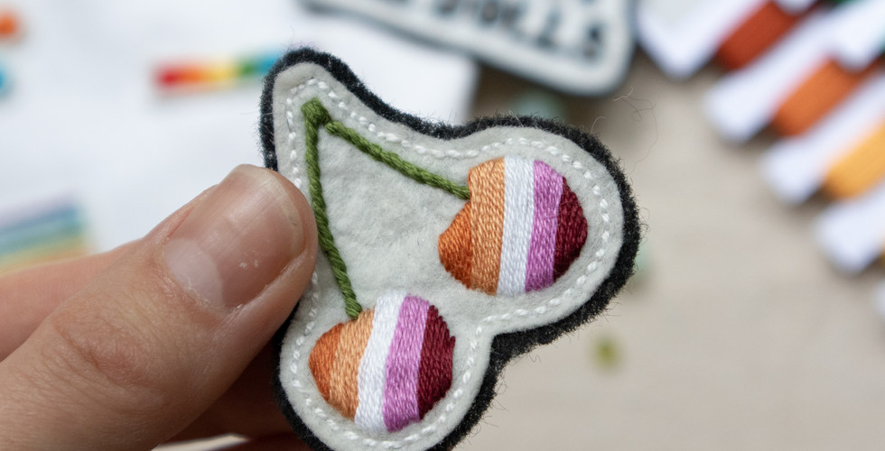 A hand embroidered patches in the shape of cherries, with the lesbian pride coloured stripes. This is a DIY embroidery patch that is being made at a queer craft workshop in London.