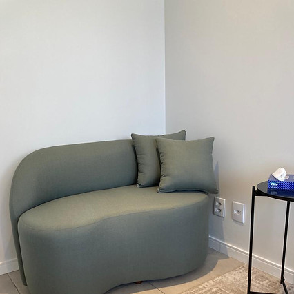 Um sofá pequeno verde musgo encostado em uma parede. Ao lado há uma mesa com lencinhos.