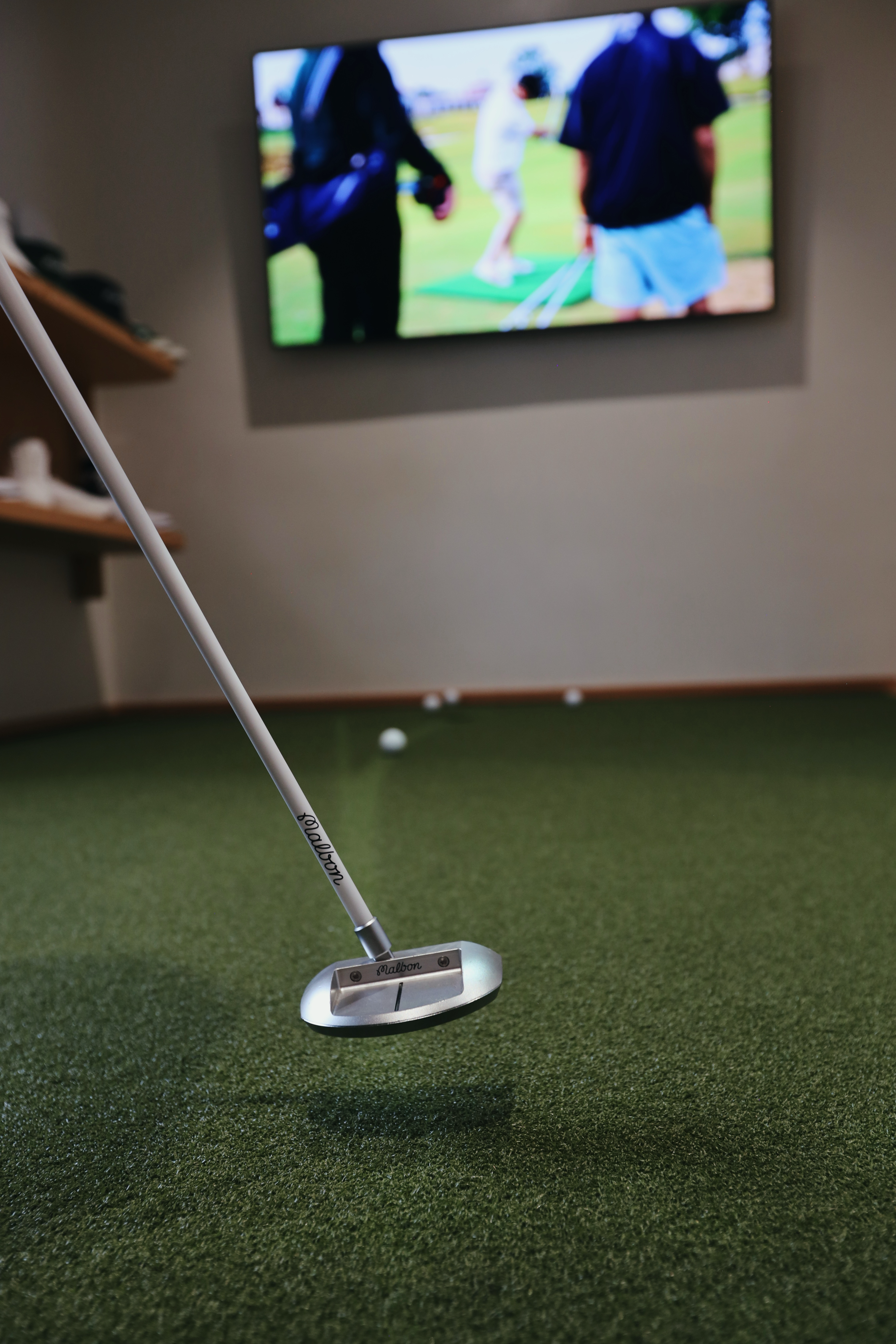 A Malbon Golf putter with a white shaft and a silver head hovering over a green putting mat, with a television screen showing people on a golf course in the background.