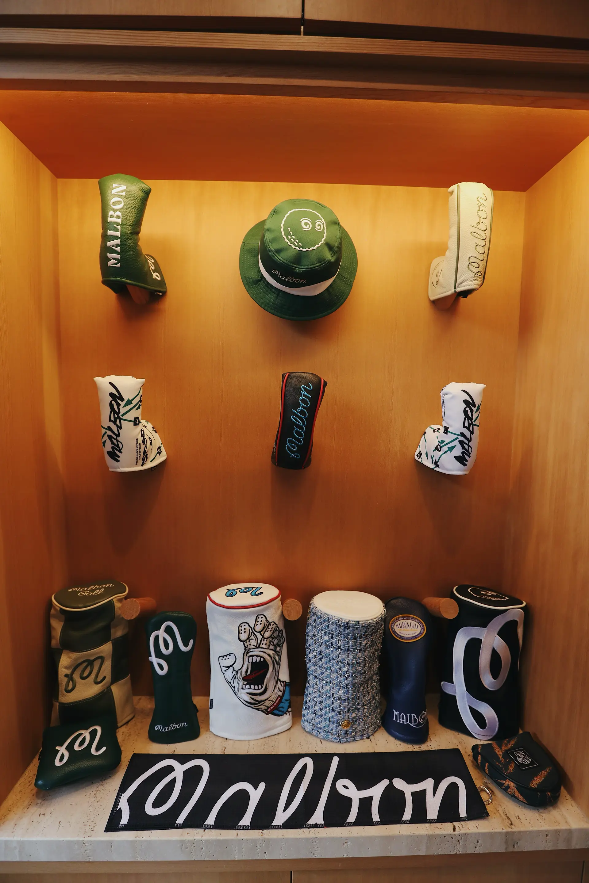 A wall display featuring a collection of Malbon Golf club headcovers, including two hanging on the back wall and a row of five on a shelf below. The collection includes a variety of designs and colors.