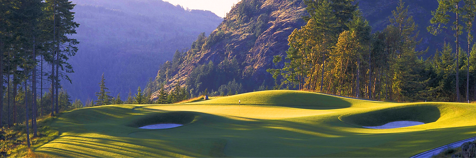 A close-up view of Bear Mountain Golf & Tennis Resort, showcasing a lush green fairway bordered by white sand traps. Towering forested hills rise sharply in the background, with the nearby mountain looming so close it feels within reach, creating a walled-in, majestic atmosphere. The course offers a stunning blend of nature’s beauty and the challenge of its dramatic surroundings.