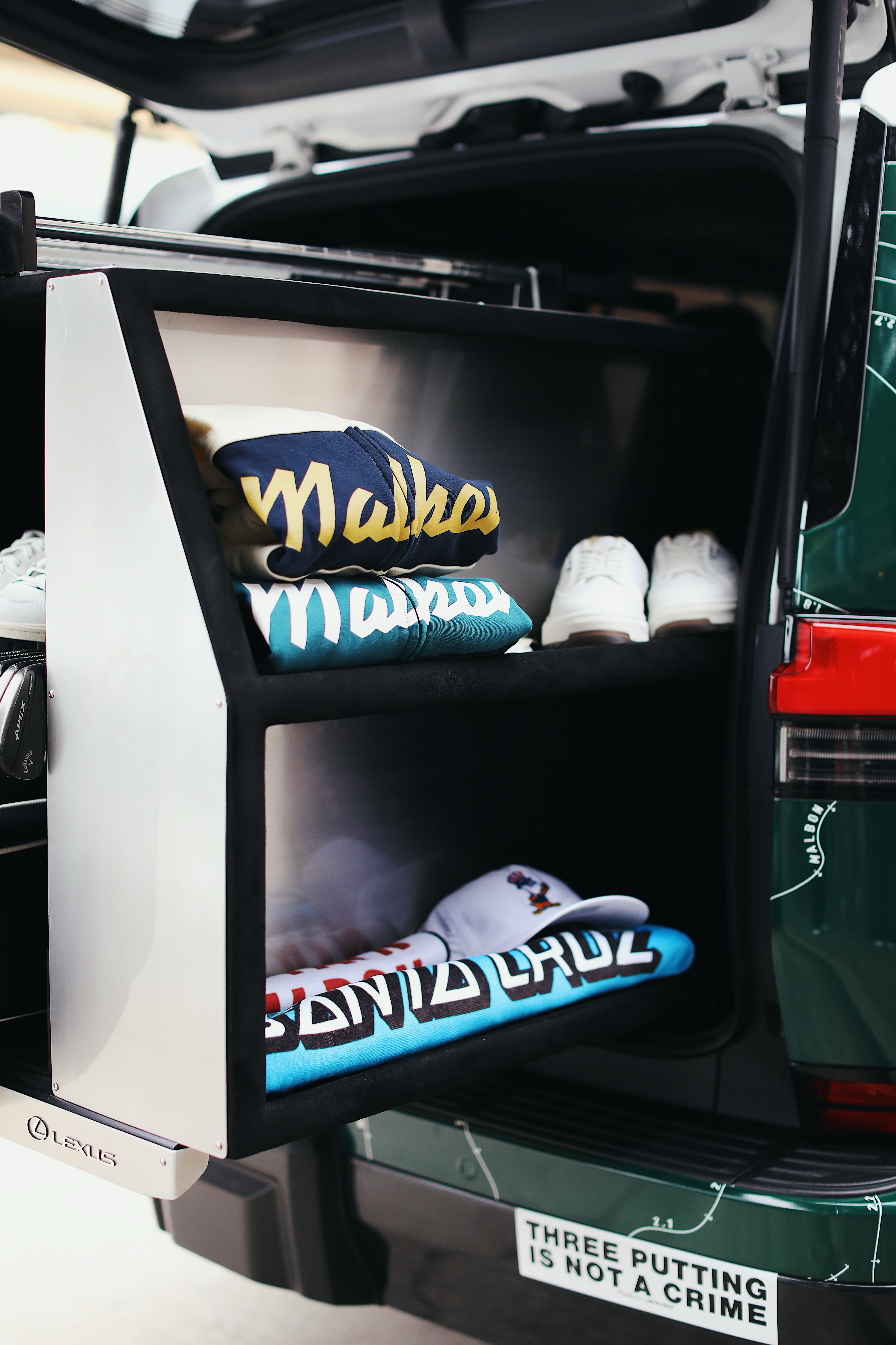 A built-in storage unit in the back of a vehicle, containing stacked Malbon Golf apparel, white sneakers, and a Santa Cruz hat. A bumper sticker below reads "THREE PUTTING IS NOT A CRIME."