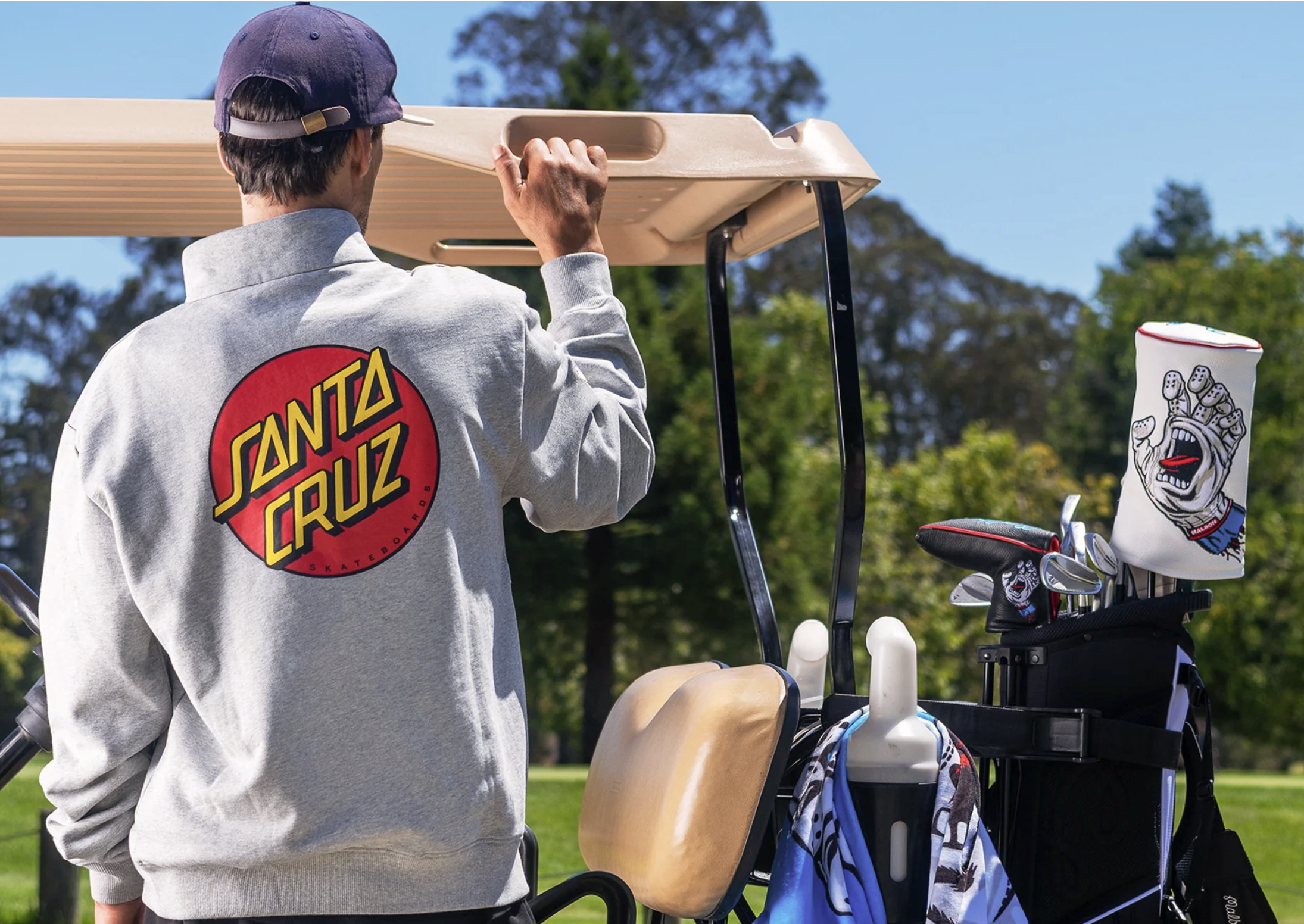 A person from behind wearing a grey sweatshirt with a large Santa Cruz logo on the back, standing next to a golf cart and golf bag. The bag holds a club headcover from the Malbon Golf x Santa Cruz collaboration with a screaming hand graphic.