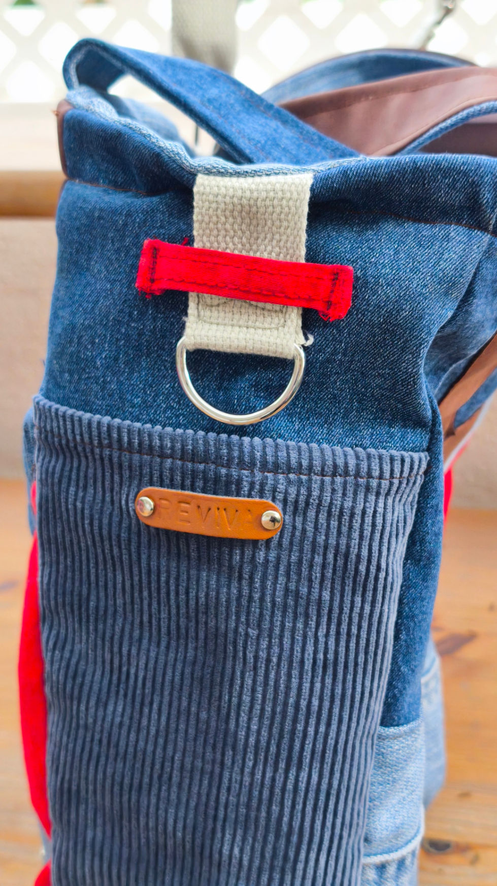 Tote bag, made from recycled denim in various shades of blue and a touch of red. Featuring a V-shaped faux leather patch and a denim and caramel-colored leather handle.