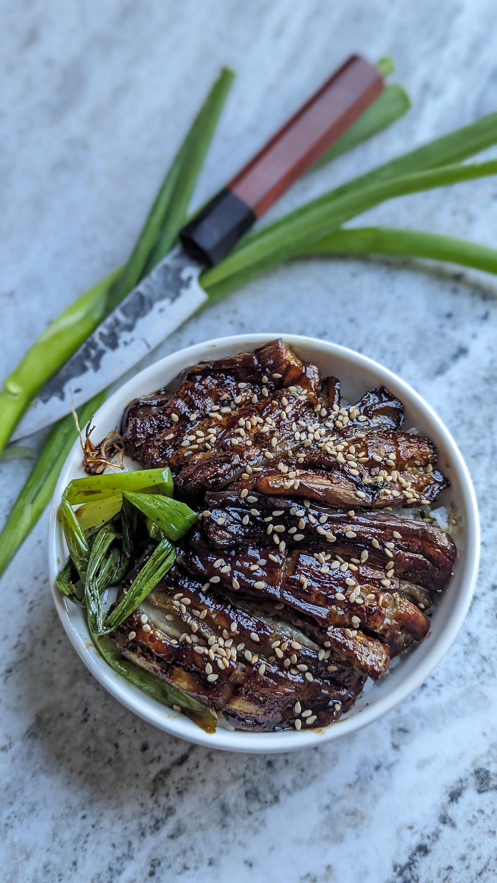 Bowl of glossy sesame-topped braised eggplant with green onions on a stone surface, beside a Forge To Table knife.