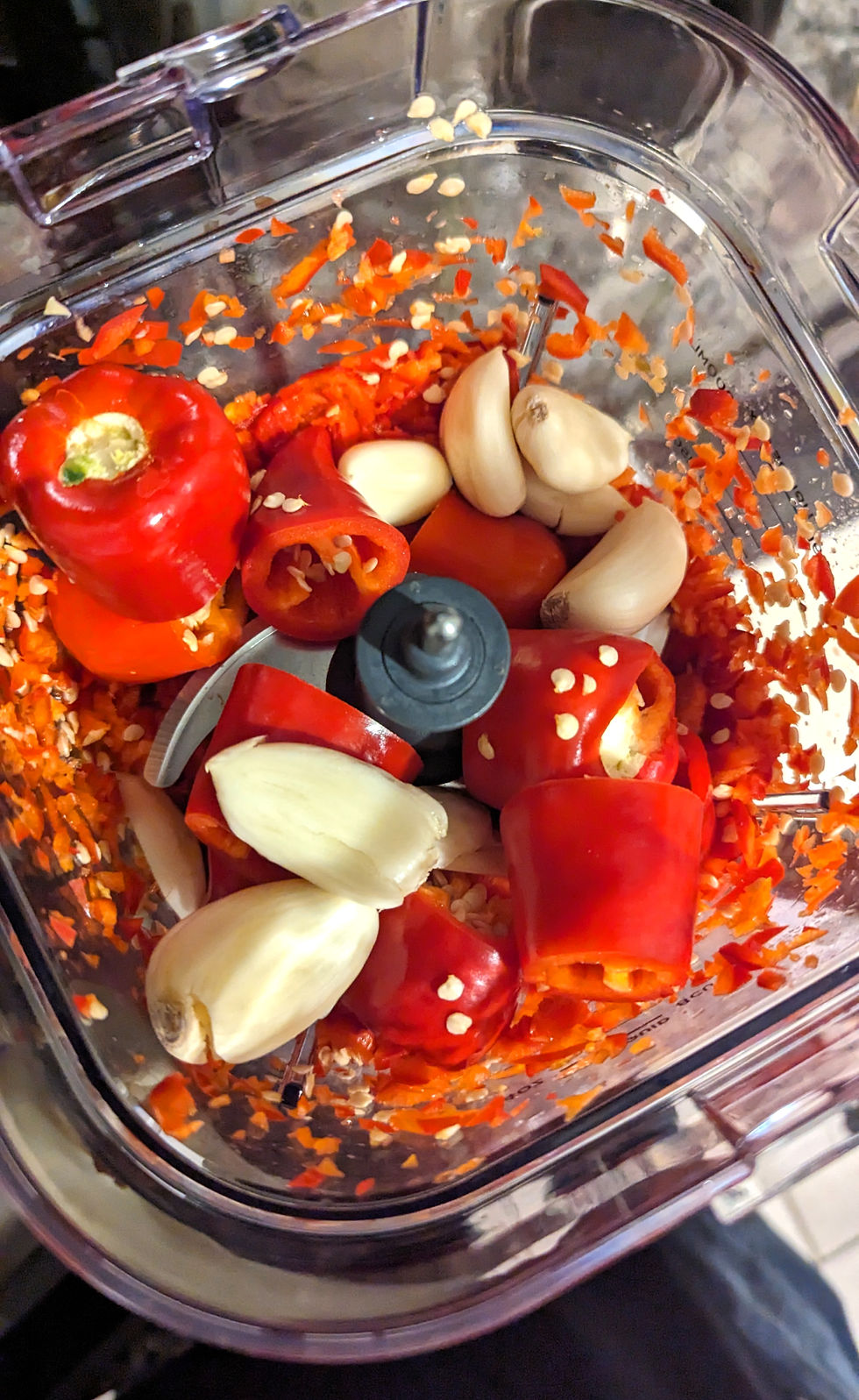 Red peppers and garlic cloves in a food processor being chopped. Bright red and white contrast against transparent container. Kitchen setting.