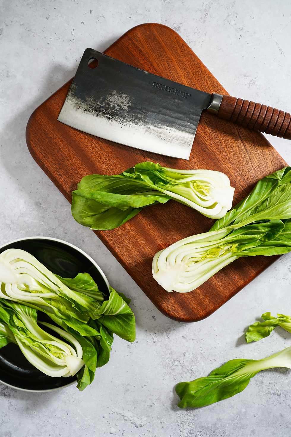 Chopped bok choy on a wooden board with a Forge To Table Classic Cleaver, beside a black bowl on a gray countertop. Green and white colors dominate the scene.