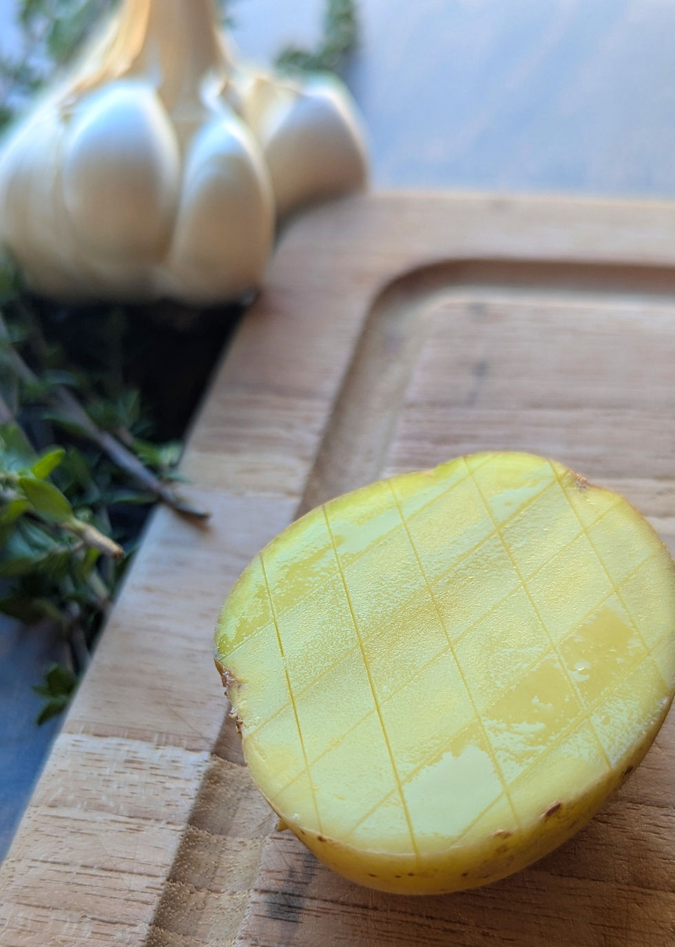 Close-up of a halved potato with a grid pattern on a wooden board, garlic and herbs in background, soft natural light.