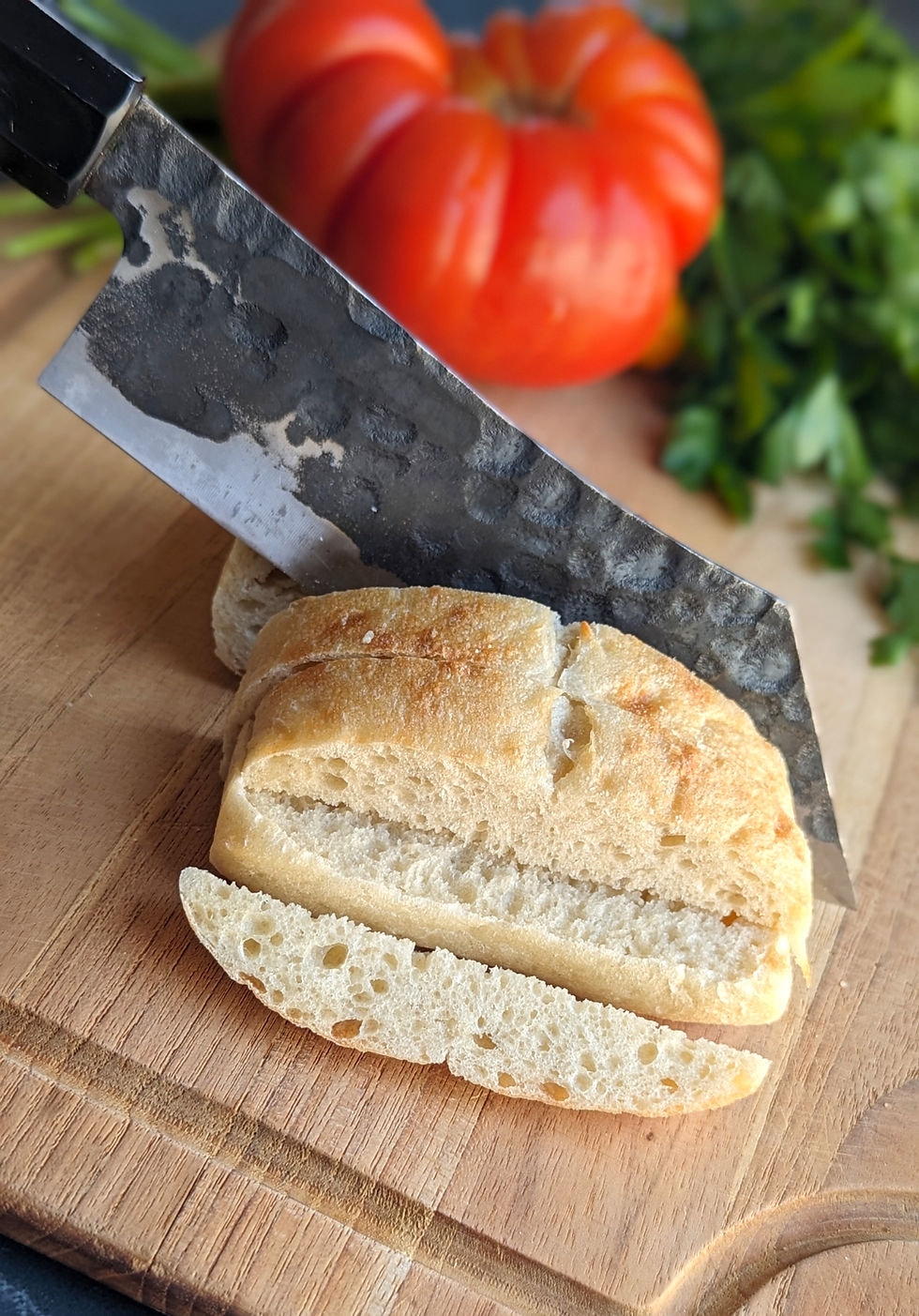 Rustic bread being sliced with a large knife on a wooden board. Background shows a red tomato and green herbs, creating a fresh mood.