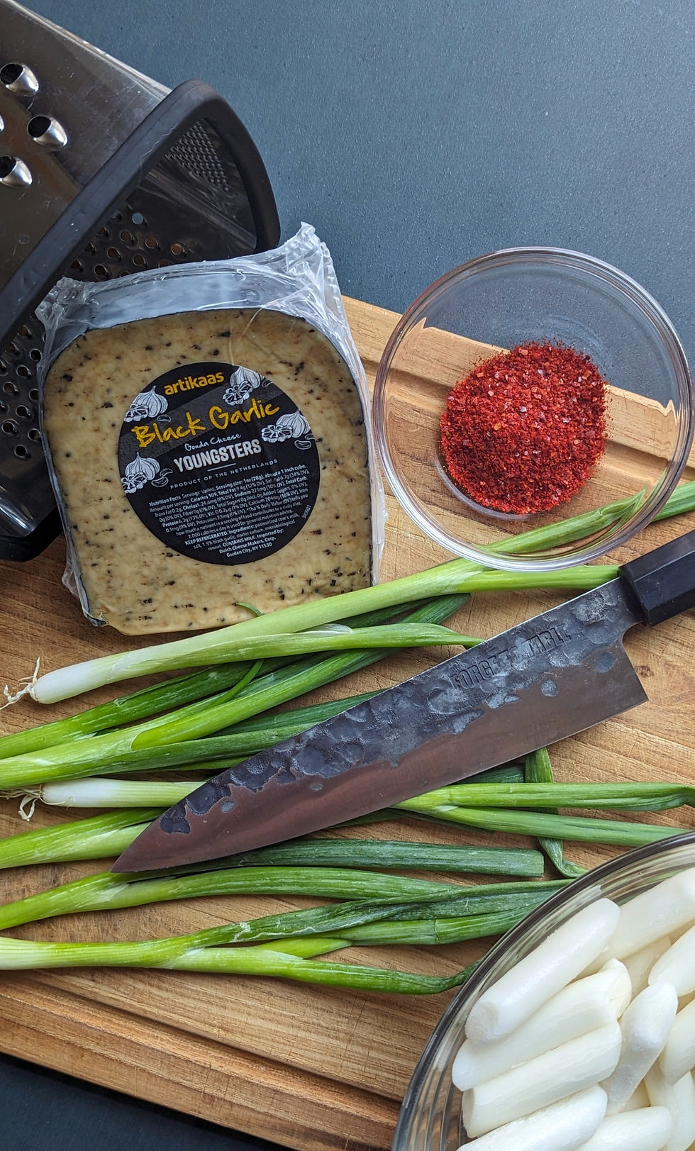 Cheese labeled "Black Garlic," green onions, a knife, red spice in a bowl on a wooden board, with a grater in the corner.
