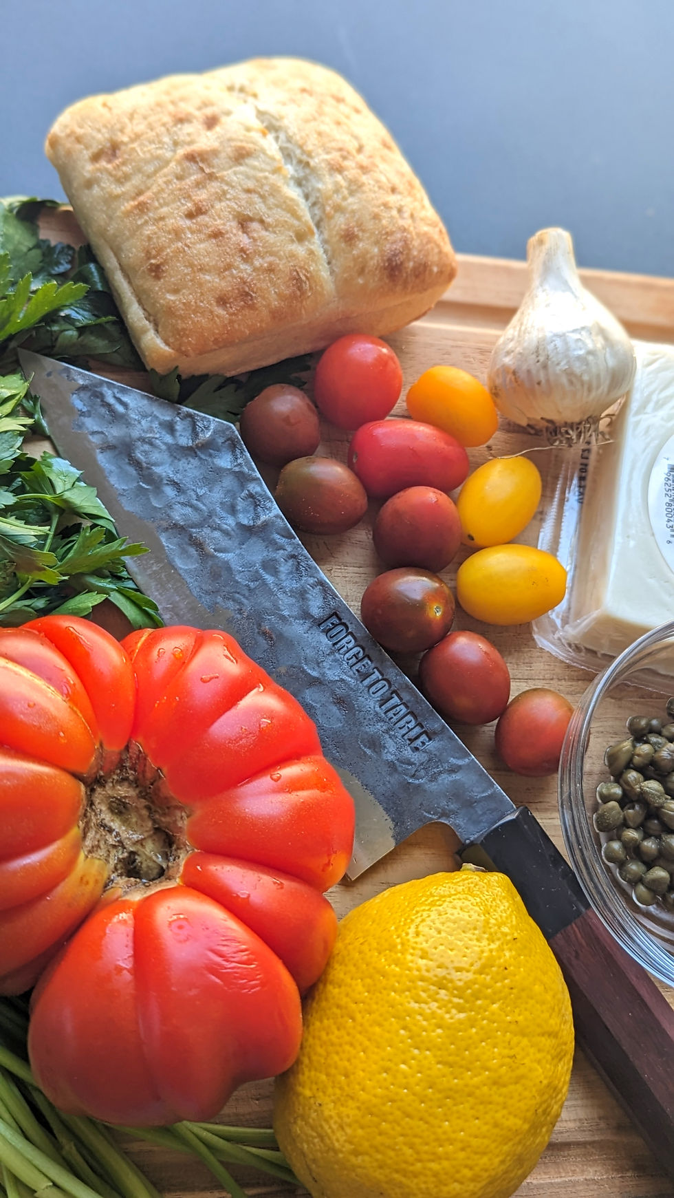 Knife on a wooden board with bread, heirloom tomato, cherry tomatoes, lemon, garlic, parsley, feta, and capers. Rustic kitchen setup.