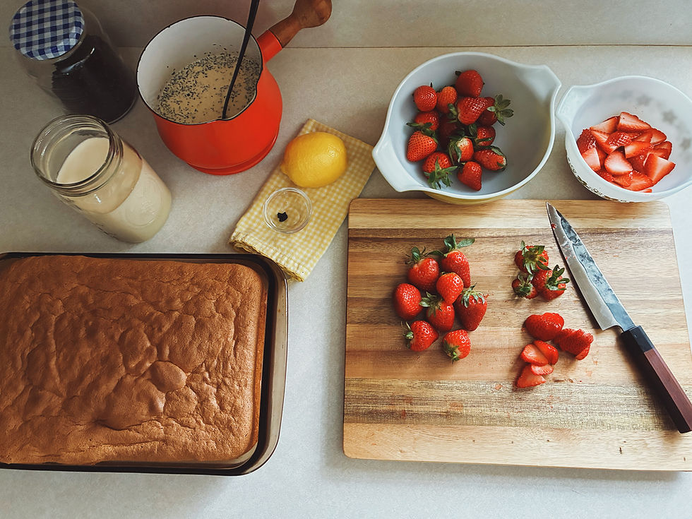 Cut strawberries on a wooden board, with a Forge To Table knife, cake, milk jar, red pot, lemon, and two bowls of strawberries on a kitchen counter.