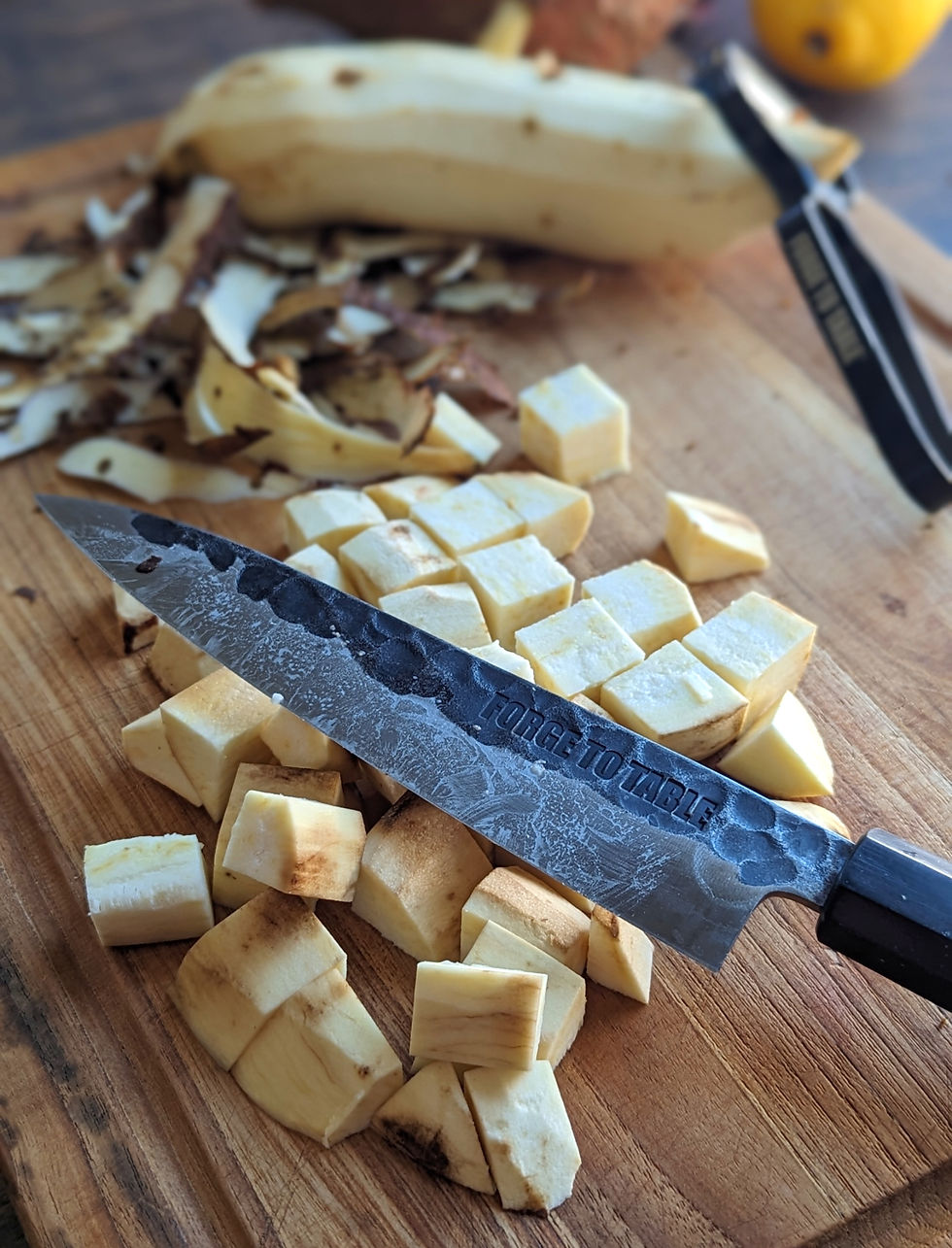 Knife with "FORGE TO TABLE" text on blade rests on chopped yam pieces. Unpeeled yam and peels on wooden board in bright kitchen setting.