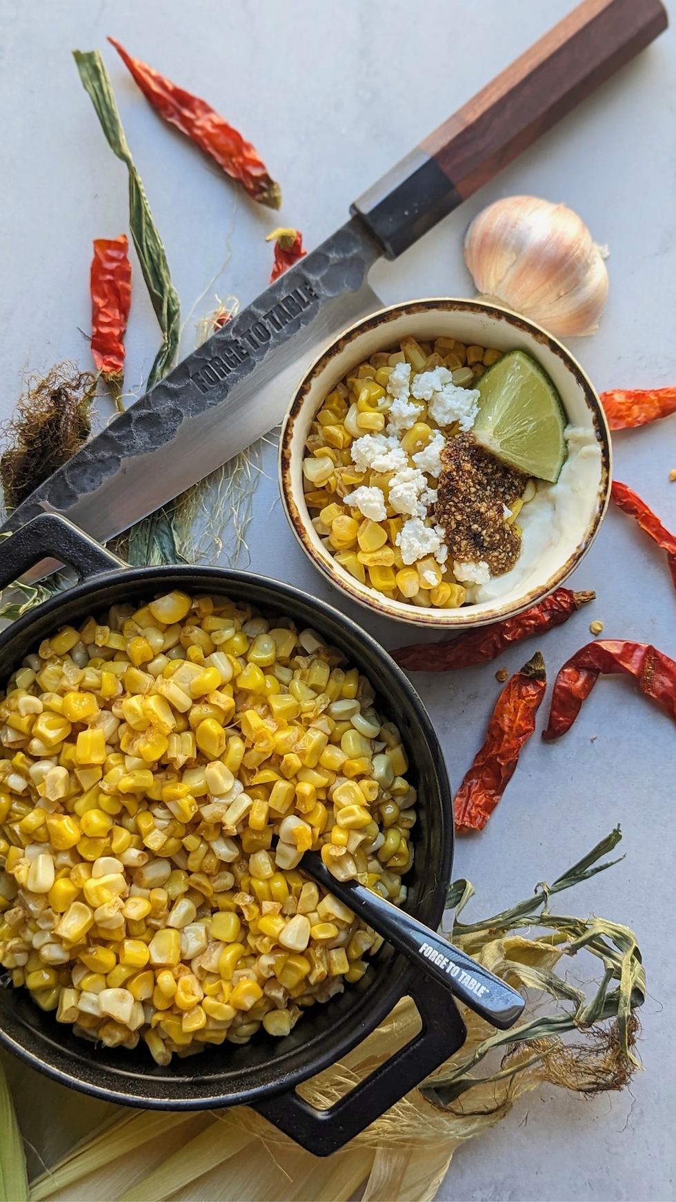 Cooked corn in a black bowl with a spoon, a knife nearby, and a smaller bowl containing corn, lime, and spices. Red chiles scattered around.