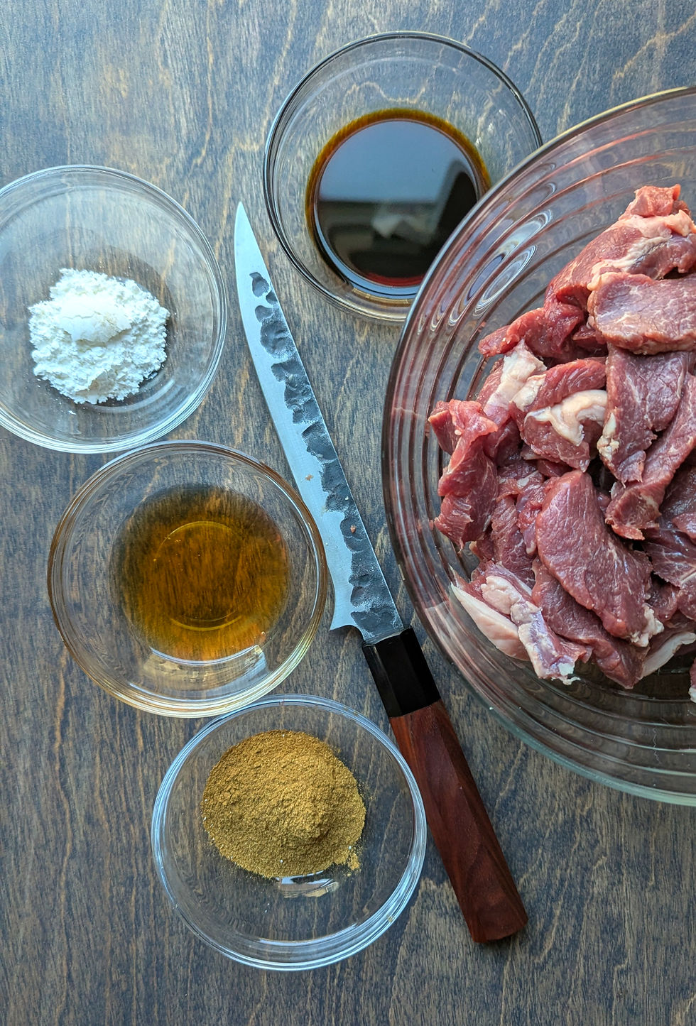 Raw beef in a bowl, surrounded by soy sauce, spices, and flour in small dishes on a wooden table. A knife rests nearby.