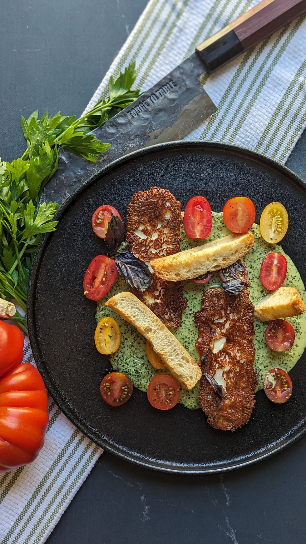 Grilled cheese and sliced cherry tomatoes on a green sauce, garnished with parsley, served on a black plate with a knife labeled "Forge to Table."