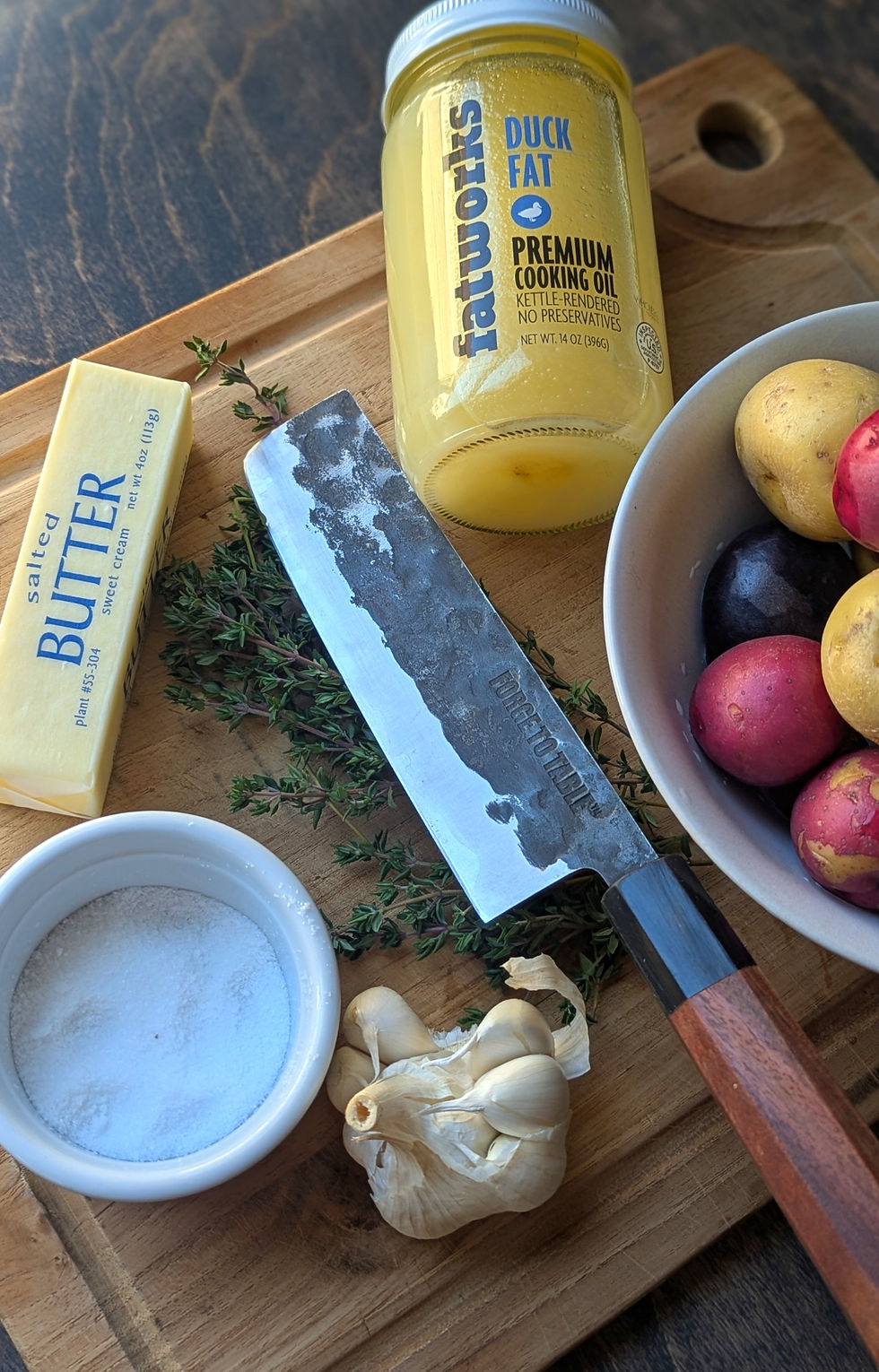 Forge To Table Nakiri knife, garlic, salt, butter, duck fat, and colorful potatoes on a wooden board. Fresh thyme adds a rustic touch.
