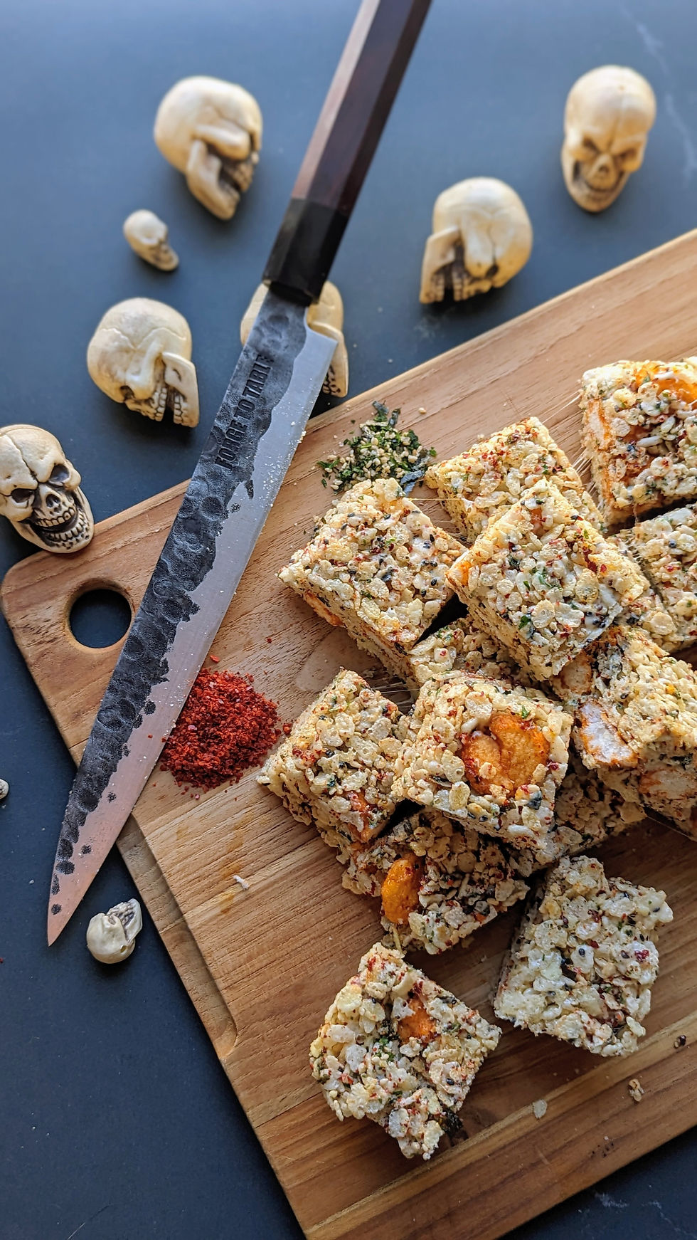 Rice cereal snack squares on a wooden board with a chef’s knife reading FORGE TO TABLE, surrounded by small skull decorations.