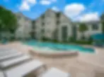 Apartment complex with a pool, surrounded by lounge chairs. The building is gray with many windows. Bright blue sky and clouds above.