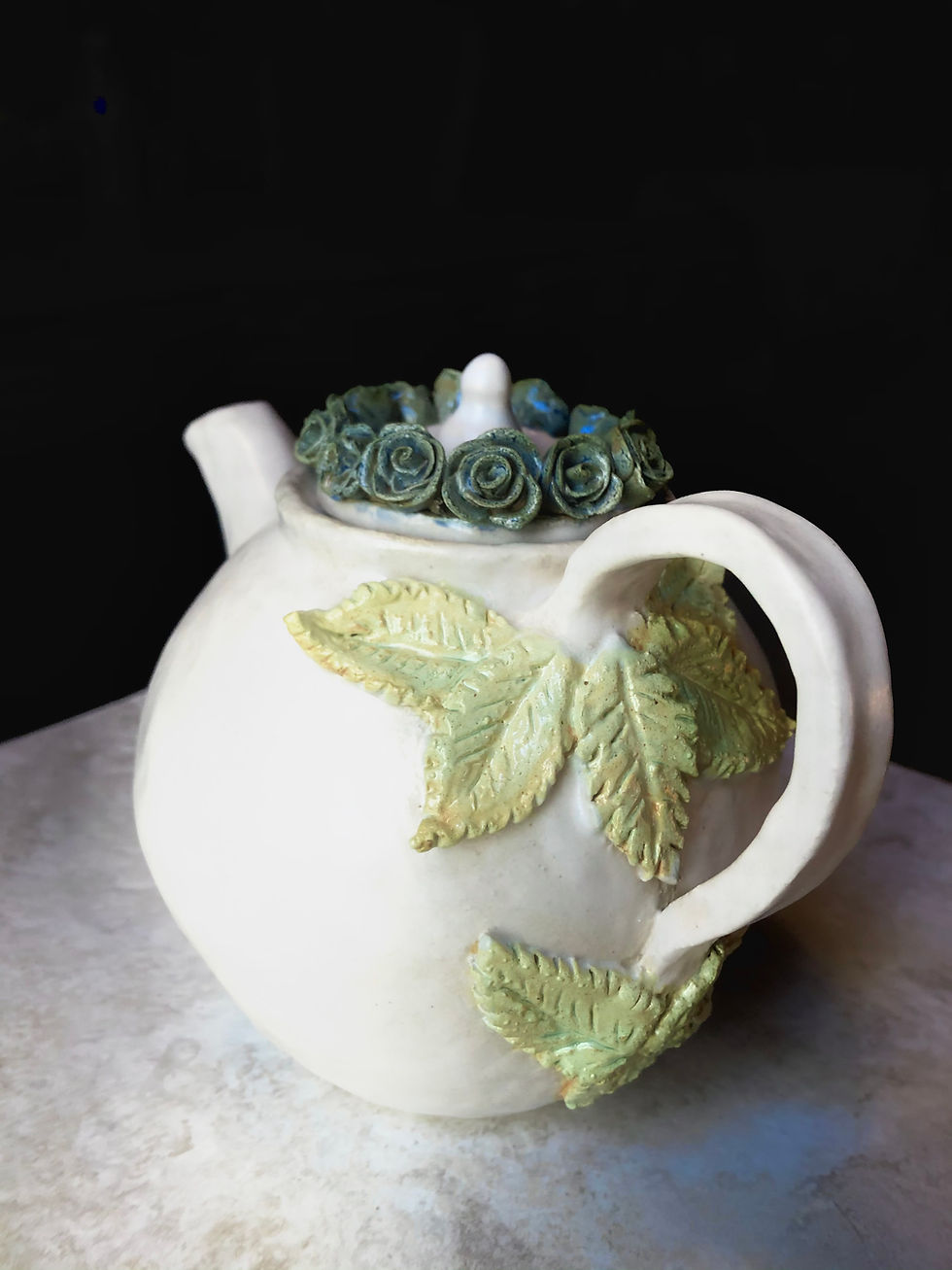 White ceramic teapot withe blue rose details on the lid and green leaves around the handle.