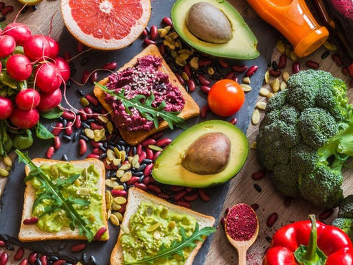 Bunte Auswahl pflanzlicher Lebensmittel auf dunklem Hintergrund: Avocadohälften, Brokkoli, Trauben, Nüsse, Samen und belegte Brote, appetitlich angerichtet