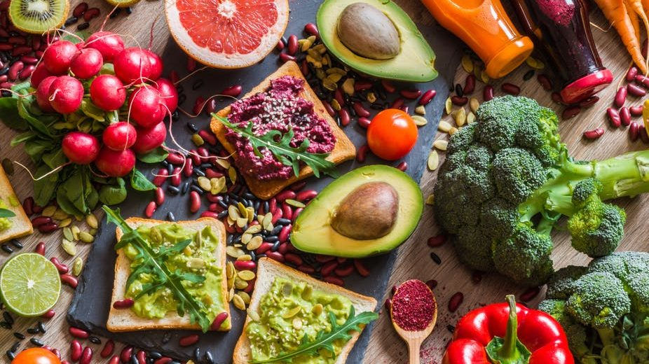 Bunte Auswahl pflanzlicher Lebensmittel auf dunklem Hintergrund: Avocadohälften, Brokkoli, Trauben, Nüsse, Samen und belegte Brote, appetitlich angerichtet